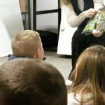 Na zdjęciu widać fragment zajęć czytelniczych prowadzonych dla dzieci w bibliotece.

Na pierwszym planie znajdują się dzieci siedzące blisko siebie na podłodze, zwrócone w stronę osoby prowadzącej. Ich postawa wskazuje na skupienie i zainteresowanie – słuchają i obserwują to, co dzieje się przed nimi.

Prowadząca siedzi na krześle i trzyma w rękach otwartą, bogato ilustrowaną książkę. Jedną ręką wskazuje konkretny element na stronie, co sugeruje, że omawia szczegóły ilustracji lub treści. Książka ma kolorowe, przyrodnicze ilustracje – widoczne są rośliny i drobne elementy natury.

W tle widać fragmenty regałów z książkami oraz wyposażenie biblioteki, co tworzy edukacyjne otoczenie. Cała scena przedstawia spokojne, angażujące zajęcia rozwijające czytelnictwo, koncentrację oraz umiejętność obserwacji u dzieci.