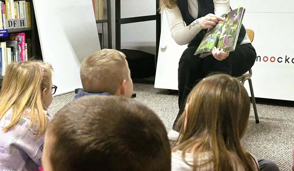 Na zdjęciu widać fragment zajęć czytelniczych prowadzonych dla dzieci w bibliotece.

Na pierwszym planie znajdują się dzieci siedzące blisko siebie na podłodze, zwrócone w stronę osoby prowadzącej. Ich postawa wskazuje na skupienie i zainteresowanie – słuchają i obserwują to, co dzieje się przed nimi.

Prowadząca siedzi na krześle i trzyma w rękach otwartą, bogato ilustrowaną książkę. Jedną ręką wskazuje konkretny element na stronie, co sugeruje, że omawia szczegóły ilustracji lub treści. Książka ma kolorowe, przyrodnicze ilustracje – widoczne są rośliny i drobne elementy natury.

W tle widać fragmenty regałów z książkami oraz wyposażenie biblioteki, co tworzy edukacyjne otoczenie. Cała scena przedstawia spokojne, angażujące zajęcia rozwijające czytelnictwo, koncentrację oraz umiejętność obserwacji u dzieci.