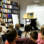 Na zdjęciu przedstawiono zajęcia edukacyjne dla dzieci odbywające się w bibliotece.

Na pierwszym planie widać grupę dzieci siedzących na podłodze, zwróconych w stronę prowadzącego. Dzieci są skupione i zaciekawione – ich uwaga koncentruje się na przedstawieniu.

Przed nimi, przy stoliku, osoba prowadząca prezentuje teatrzyk kamishibai – drewnianą skrzynkę z ilustracją widoczną w okienku. Na obrazku znajduje się kolorowa scena z postacią dziewczynki i elementami przyrody, co sugeruje opowieść o charakterze bajkowym lub edukacyjnym.

W tle widoczne są regały pełne książek oraz sprzęt multimedialny, co jednoznacznie wskazuje na przestrzeń biblioteczną. Obok stoi tablica lub flipchart, który może być wykorzystywany podczas zajęć.

Cała scena oddaje atmosferę aktywnego czytelnictwa i edukacji – dzieci uczestniczą w angażującej formie pracy z książką i obrazem, rozwijając wyobraźnię oraz umiejętność słuchania.