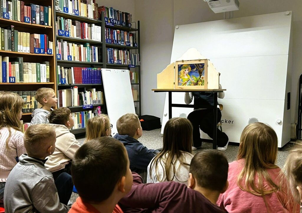 Na zdjęciu przedstawiono zajęcia edukacyjne dla dzieci odbywające się w bibliotece.

Na pierwszym planie widać grupę dzieci siedzących na podłodze, zwróconych w stronę prowadzącego. Dzieci są skupione i zaciekawione – ich uwaga koncentruje się na przedstawieniu.

Przed nimi, przy stoliku, osoba prowadząca prezentuje teatrzyk kamishibai – drewnianą skrzynkę z ilustracją widoczną w okienku. Na obrazku znajduje się kolorowa scena z postacią dziewczynki i elementami przyrody, co sugeruje opowieść o charakterze bajkowym lub edukacyjnym.

W tle widoczne są regały pełne książek oraz sprzęt multimedialny, co jednoznacznie wskazuje na przestrzeń biblioteczną. Obok stoi tablica lub flipchart, który może być wykorzystywany podczas zajęć.

Cała scena oddaje atmosferę aktywnego czytelnictwa i edukacji – dzieci uczestniczą w angażującej formie pracy z książką i obrazem, rozwijając wyobraźnię oraz umiejętność słuchania.