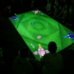 Na zdjęciu widać grupę dzieci uczestniczących w zajęciach w bibliotece, siedzących i klęczących na dywanie pomiędzy regałami z książkami. Na podłodze wyświetlana jest kolorowa projekcja świetlna, która przyciąga uwagę dzieci i staje się centrum wspólnej aktywności.

Kilka dzieci aktywnie wchodzi w interakcję z obrazem: dotykają światła, wskazują elementy, przesuwają się po dywanie, jakby „wchodziły” w wyświetlaną scenę. Inne siedzą wokół w kręgu, obserwując z zainteresowaniem i czekając na swoją kolej. Atmosfera jest skupiona, ale jednocześnie swobodna i pełna ciekawości.
Zdjęcie pokazuje:
nowoczesne, angażujące formy pracy z dziećmi w bibliotece,
naukę poprzez zabawę i doświadczenie,
bibliotekę jako przestrzeń interaktywną i przyjazną najmłodszym.