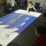 Na zdjęciu widać grupę dzieci uczestniczących w zajęciach w bibliotece, siedzących i klęczących na dywanie pomiędzy regałami z książkami. Na podłodze wyświetlana jest kolorowa projekcja świetlna, która przyciąga uwagę dzieci i staje się centrum wspólnej aktywności.

Dziewczynka aktywnie wchodzi w interakcję z obrazem: dotykają światła, wskazują elementy, przesuwają się po dywanie, jakby „wchodziły” w wyświetlaną scenę. Inne dzieci siedzą wokół w kręgu, obserwując z zainteresowaniem i czekając na swoją kolej. Atmosfera jest skupiona, ale jednocześnie swobodna i pełna ciekawości.
Zdjęcie pokazuje:
nowoczesne, angażujące formy pracy z dziećmi w bibliotece,
naukę poprzez zabawę i doświadczenie,
bibliotekę jako przestrzeń interaktywną i przyjazną najmłodszym.