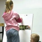 Na zdjęciu widać fragment zajęć edukacyjnych dla dzieci.

Na pierwszym planie stoi dziewczynka ubrana w różową bluzę i dżinsy, która pracuje przy tablicy lub flipcharcie. Przypina do niej obrazki – widoczne są fotografie przedstawiające elementy przyrody, m.in. kwiaty (prawdopodobnie konwalie) oraz ptaki. Obrazki są przymocowane za pomocą kolorowych magnesów.

Obok, nieco niżej, siedzi chłopiec, który uważnie obserwuje działania koleżanki. Jego postawa wskazuje na zainteresowanie i zaangażowanie w zadanie.

Tło jest proste i jasne, co kieruje uwagę na aktywność dzieci.