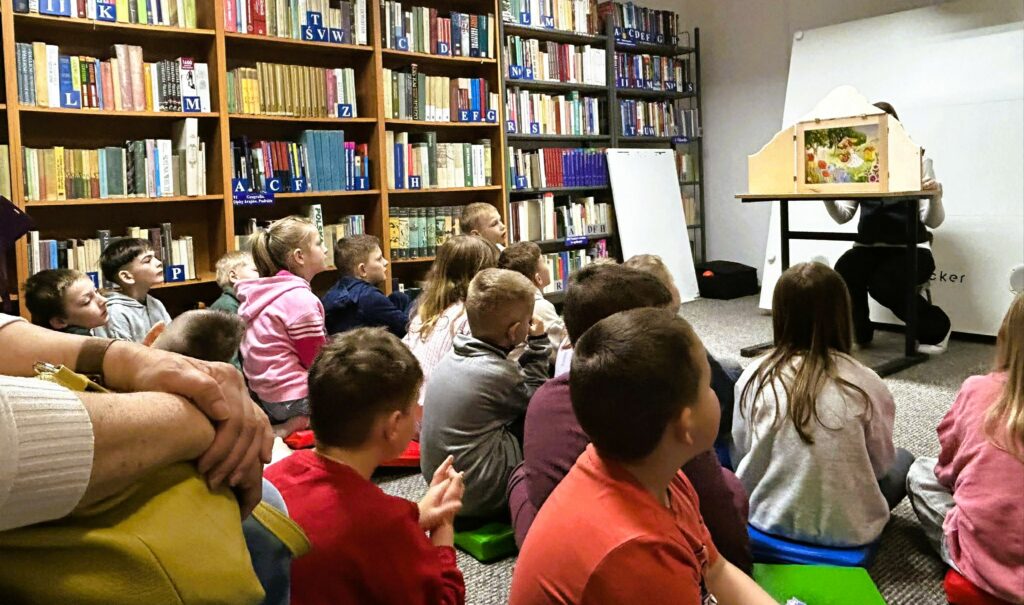 Na zdjęciu przedstawiono zajęcia edukacyjne dla dzieci odbywające się w bibliotece.

Na pierwszym planie widać grupę dzieci siedzących na podłodze, zwróconych w stronę prowadzącego. Dzieci są skupione i zaciekawione – ich uwaga koncentruje się na przedstawieniu.

Przed nimi, przy stoliku, osoba prowadząca prezentuje teatrzyk kamishibai – drewnianą skrzynkę z ilustracją widoczną w okienku. Na obrazku znajduje się kolorowa scena z postacią dziewczynki i elementami przyrody, co sugeruje opowieść o charakterze bajkowym lub edukacyjnym.

W tle widoczne są regały pełne książek oraz sprzęt multimedialny, co jednoznacznie wskazuje na przestrzeń biblioteczną. Obok stoi tablica lub flipchart, który może być wykorzystywany podczas zajęć.

Cała scena oddaje atmosferę aktywnego czytelnictwa i edukacji – dzieci uczestniczą w angażującej formie pracy z książką i obrazem, rozwijając wyobraźnię oraz umiejętność słuchania.