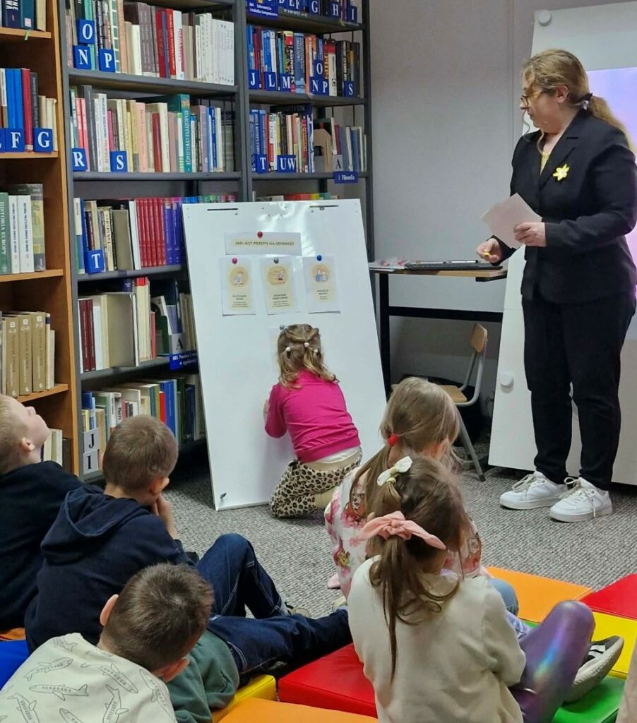 Na zdjęciu widoczne są zajęcia edukacyjne prowadzone w bibliotece.

Na pierwszym planie siedzi grupa dzieci na kolorowych matach, uważnie obserwujących koleżankę, która podeszła do tablicy i przypina kartę lub wykonuje zadanie. Obok stoi prowadząca – kobieta z przypiętym żółtym żonkilem, co wskazuje na udział w Akcji Żonkile.

Na tablicy znajdują się ilustracje i krótkie opisy – prawdopodobnie dotyczące postaw lub zachowań związanych z odwagą (widoczny napis sugeruje temat „Jaki jest przepis na odwagę?”). Dzieci aktywnie uczestniczą w zajęciach, biorąc udział w ćwiczeniu polegającym na wyborze lub dopasowaniu odpowiednich elementów.

W tle widać regały wypełnione książkami, co podkreśla, że zajęcia odbywają się w przestrzeni bibliotecznej.

Zdjęcie ukazuje angażującą formę nauki poprzez działanie i współpracę, związaną z tematyką wartości takich jak odwaga i empatia, nawiązującą do pamięci o Powstanie w getcie warszawskim.