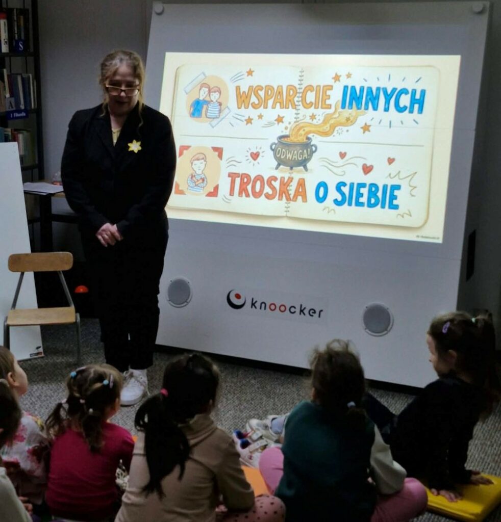 Na zdjęciu widoczna jest scena z zajęć edukacyjnych prowadzonych w bibliotece.

Na pierwszym planie siedzi grupa dzieci, które uważnie słuchają prowadzącej stojącej obok tablicy multimedialnej. Kobieta ma przypięty żółty żonkil – symbol udziału w Akcji Żonkile.

Na ekranie wyświetlona jest kolorowa ilustracja z hasłami: „Wsparcie innych” oraz „Troska o siebie”, a w centrum znajduje się słowo „odwaga”. Grafika sugeruje, że tematem zajęć są wartości takie jak empatia, dbanie o siebie i pomaganie innym.

Dzieci siedzą spokojnie, skupione na przekazie, co wskazuje na rozmowę lub wspólne omawianie przedstawionych treści. W tle widać elementy wyposażenia biblioteki.

Zdjęcie ukazuje edukację poprzez rozmowę i refleksję, najprawdopodobniej w kontekście zajęć związanych z pamięcią o Powstanie w getcie warszawskim oraz wartościami takimi jak odwaga i solidarność.