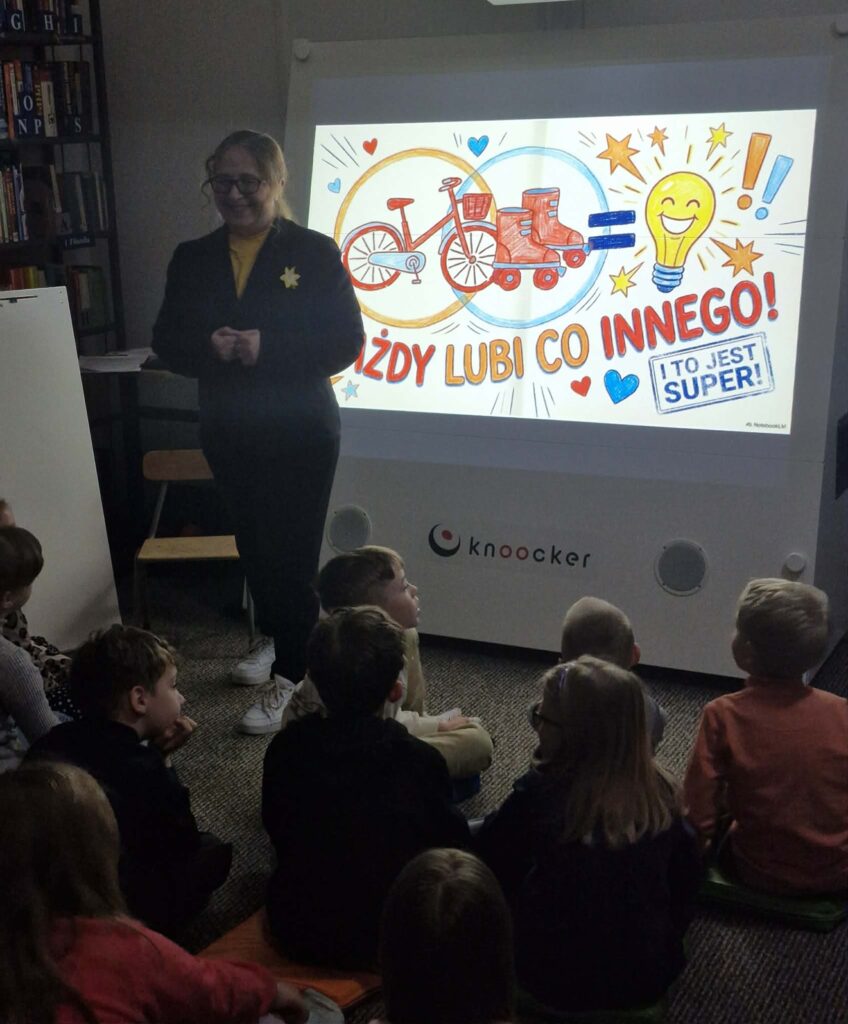 Na zdjęciu przedstawiono zajęcia edukacyjne prowadzone w bibliotece dla grupy dzieci.

Na pierwszym planie dzieci siedzą na podłodze i uważnie słuchają prowadzącej. Kobieta stoi obok dużego ekranu interaktywnego (marki „knoocker”) i uśmiecha się, prowadząc rozmowę lub wyjaśniając temat. Ma przypięty żółty żonkil, co wskazuje na udział w działaniach związanych z pamięcią historyczną.

Na ekranie wyświetlona jest kolorowa grafika z hasłem: „Każdy lubi co innego! I to jest super!”. Ilustracje (rower, rolki, żarówka, serca) podkreślają temat różnorodności, indywidualnych zainteresowań i akceptacji innych.

W tle widać regały z książkami, co potwierdza, że zajęcia odbywają się w bibliotece. Cała scena pokazuje aktywną, angażującą formę pracy z dziećmi, łączącą edukację z rozmową o wartościach takich jak tolerancja, akceptacja i szacunek dla różnic między ludźmi.