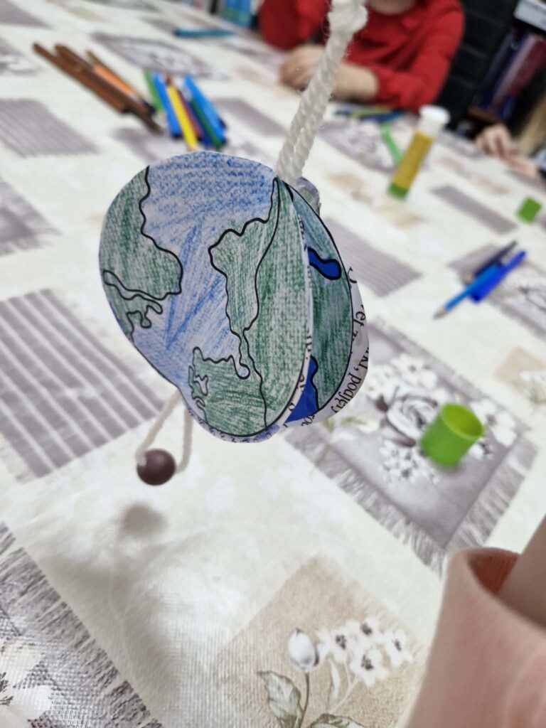 Na zdjęciu widoczna jest praca plastyczna wykonana z papieru – niewielka zawieszka ozdobione rysunkiem przypominającym kulę ziemską. Na jego powierzchni narysowano kontynenty w kolorze zielonym oraz żółtym, otoczone niebieskimi oceanami.

Praca została wykonana ręcznie, widać charakterystyczne ślady kolorowania kredkami lub flamastrami oraz lekko nieregularne kształty. Całość ma formę przestrzenną (3D).

Zawieszka leży na stole przykrytym wzorzystym obrusem. Praca jest związana z zajęciami o tematyce ekologicznej rozwijającymi kreatywność i wiedzę o świecie.