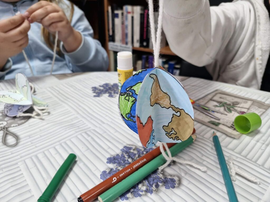 Na zdjęciu widoczna jest praca plastyczna wykonana z papieru – niewielka zawieszka ozdobione rysunkiem przypominającym kulę ziemską. Na jego powierzchni narysowano kontynenty w kolorze zielonym oraz żółtym, otoczone niebieskimi oceanami.

Praca została wykonana ręcznie, widać charakterystyczne ślady kolorowania kredkami lub flamastrami oraz lekko nieregularne kształty. Całość ma formę przestrzenną (3D).

Zawieszka leży na stole przykrytym wzorzystym obrusem. Praca jest związana z zajęciami o tematyce ekologicznej rozwijającymi kreatywność i wiedzę o świecie.