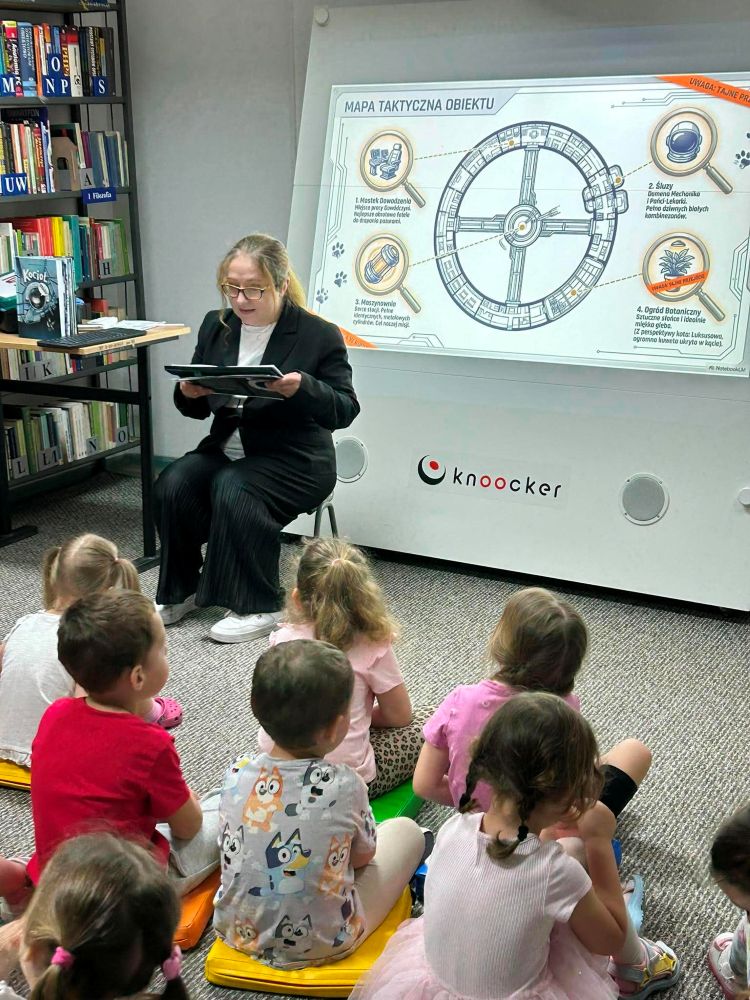 Na zdjęciu widoczne są zajęcia biblioteczno-edukacyjne dla dzieci poświęcone tematyce kosmosu i wyobraźni. Prowadząca siedzi przed grupą dzieci i czyta książkę lub fragment opowieści, angażując uczestników we wspólną podróż po świecie fantazji.
W tle wyświetlona została ilustracja przypominająca „mapę taktyczną obiektu” — futurystyczny plan stacji lub statku kosmicznego, co dodatkowo buduje atmosferę międzyplanetarnej przygody.
Dzieci siedzą na kolorowych poduszkach i z dużym zainteresowaniem słuchają opowieści. Obok widoczna jest książka Kocioł: pierwszy astromiauta, która stanowi inspirację do zajęć.
Fotografia pokazuje:
nowoczesne i kreatywne zajęcia biblioteczne,
połączenie literatury z multimediami,
rozwijanie wyobraźni i ciekawości świata,
aktywne uczestnictwo dzieci w spotkaniu edukacyjnym