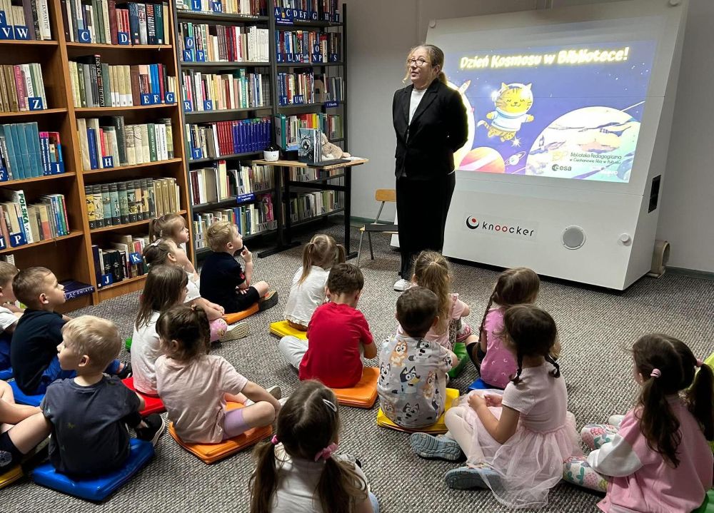 Na zdjęciu przedstawiono zajęcia edukacyjne dla dzieci odbywające się w bibliotece. Prowadząca stoi przed grupą dzieci siedzących na kolorowych poduszkach i opowiada o kosmosie, angażując uczestników rozmową oraz gestami.
W tle widoczna jest prezentacja multimedialna z napisem „Dzień Kosmosu w Bibliotece!” oraz ilustracją kota astronauty, nawiązującą do książki Kocioł: pierwszy astromiauta.
Atmosfera zajęć wydaje się bardzo przyjazna i pełna zaciekawienia. Dzieci z uwagą słuchają prowadzącej, co podkreśla edukacyjny i angażujący charakter spotkania.
Zdjęcie pokazuje:
nowoczesne wykorzystanie prezentacji multimedialnej,
aktywne metody pracy z dziećmi,
promocję czytelnictwa i nauki poprzez zabawę,
bibliotekę jako przestrzeń inspirujących spotkań edukacyjnych