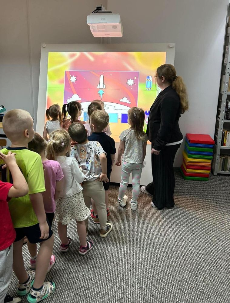 Na zdjęciu widać zajęcia edukacyjne z wykorzystaniem tablicy multimedialnej w bibliotece.
Na pierwszym planie stoi kilkoro dzieci, które podchodzą do dużego ekranu i aktywnie uczestniczą w zadaniu. Na tablicy wyświetlona jest kolorowa grafika przedstawiająca kosmos – statek kosmiczny, planety i gwiazdy. Jedno z dzieci dotyka ekranu, co sugeruje, że jest to interaktywna aplikacja lub gra edukacyjna.
Z boku stoi dorosła kobieta (prowadząca zajęcia), która obserwuje działania dzieci i wspiera ich w wykonywaniu zadania.
W tle widoczne są regały z książkami, co potwierdza, że zajęcia odbywają się w przestrzeni biblioteki. Całość przedstawia nowoczesne podejście do edukacji – łączenie tradycyjnej biblioteki z technologią oraz nauką poprzez zabawę.