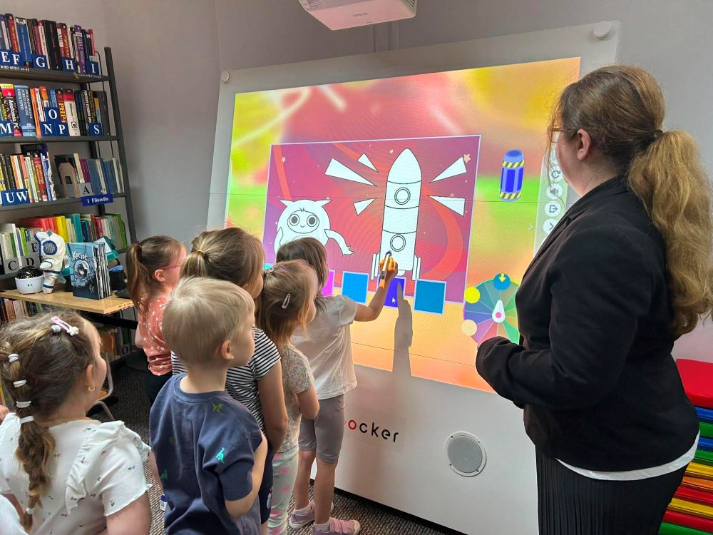 Na zdjęciu widać zajęcia edukacyjne z wykorzystaniem tablicy multimedialnej w bibliotece.
Na pierwszym planie stoi kilkoro dzieci, które podchodzą do dużego ekranu i aktywnie uczestniczą w zadaniu. Na tablicy wyświetlona jest kolorowa grafika przedstawiająca kosmos – statek kosmiczny, planety i gwiazdy. Jedno z dzieci dotyka ekranu, co sugeruje, że jest to interaktywna aplikacja lub gra edukacyjna.
Z boku stoi dorosła kobieta (prowadząca zajęcia), która obserwuje działania dzieci i wspiera ich w wykonywaniu zadania.
W tle widoczne są regały z książkami, co potwierdza, że zajęcia odbywają się w przestrzeni biblioteki. Całość przedstawia nowoczesne podejście do edukacji – łączenie tradycyjnej biblioteki z technologią oraz nauką poprzez zabawę.
