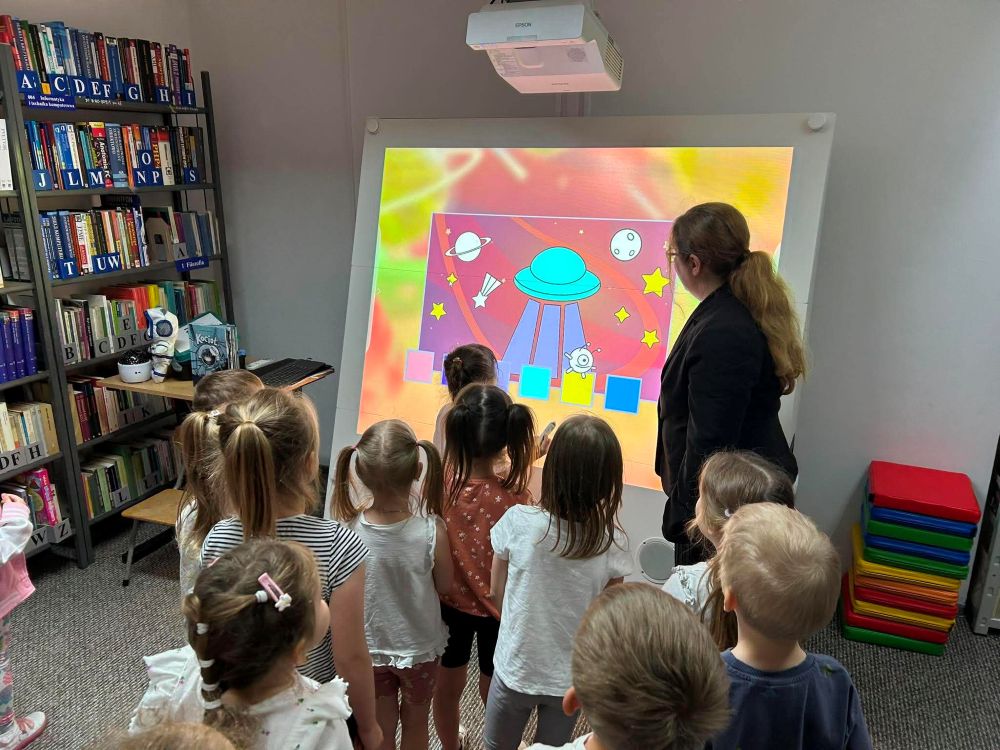 Na zdjęciu widać zajęcia edukacyjne z wykorzystaniem tablicy multimedialnej w bibliotece.
Na pierwszym planie stoi kilkoro dzieci, które podchodzą do dużego ekranu i aktywnie uczestniczą w zadaniu. Na tablicy wyświetlona jest kolorowa grafika przedstawiająca kosmos – statek kosmiczny, planety i gwiazdy. Jedno z dzieci dotyka ekranu, co sugeruje, że jest to interaktywna aplikacja lub gra edukacyjna.
Z boku stoi dorosła kobieta (prowadząca zajęcia), która obserwuje działania dzieci i wspiera ich w wykonywaniu zadania.
W tle widoczne są regały z książkami, co potwierdza, że zajęcia odbywają się w przestrzeni biblioteki. Całość przedstawia nowoczesne podejście do edukacji – łączenie tradycyjnej biblioteki z technologią oraz nauką poprzez zabawę.