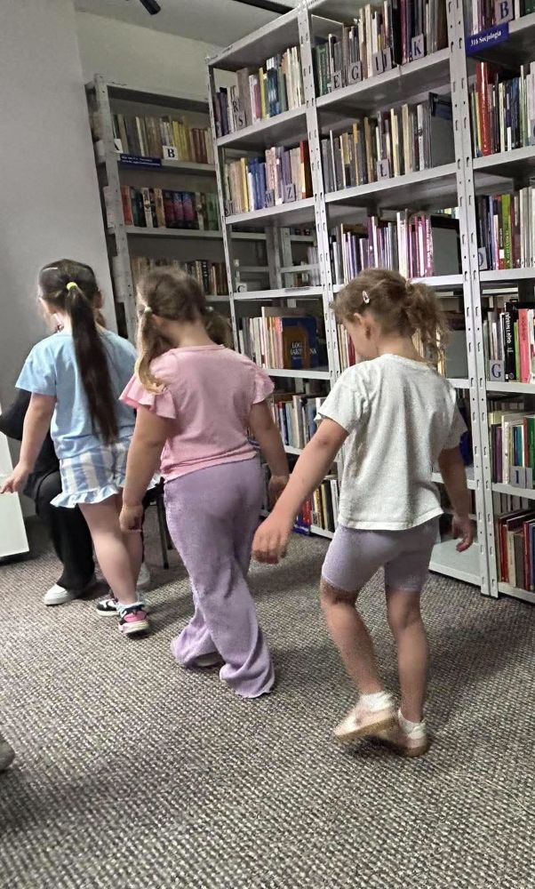 Na zdjęciu widać troje dzieci uczestniczących w zajęciach ruchowych lub integracyjnych odbywających się w przestrzeni bibliotecznej.
Dziewczynki idą jedna za drugą pomiędzy regałami z książkami, wykonując spokojne ćwiczenie ruchowe. Atmosfera zdjęcia jest swobodna i przyjazna, a aktywność wygląda na element zabawy edukacyjnej lub ćwiczeń wspierających koncentrację i współpracę w grupie.
Fotografia pokazuje:
połączenie nauki i ruchu podczas zajęć bibliotecznych,
aktywne uczestnictwo dzieci,
przyjazną przestrzeń biblioteki otwartą na najmłodszych,
rozwijanie koordynacji i integracji poprzez zabawę 🌿👣
W tle widoczne są wysokie biblioteczne regały wypełnione książkami, które tworzą kameralny i edukacyjny klimat spotkania.
