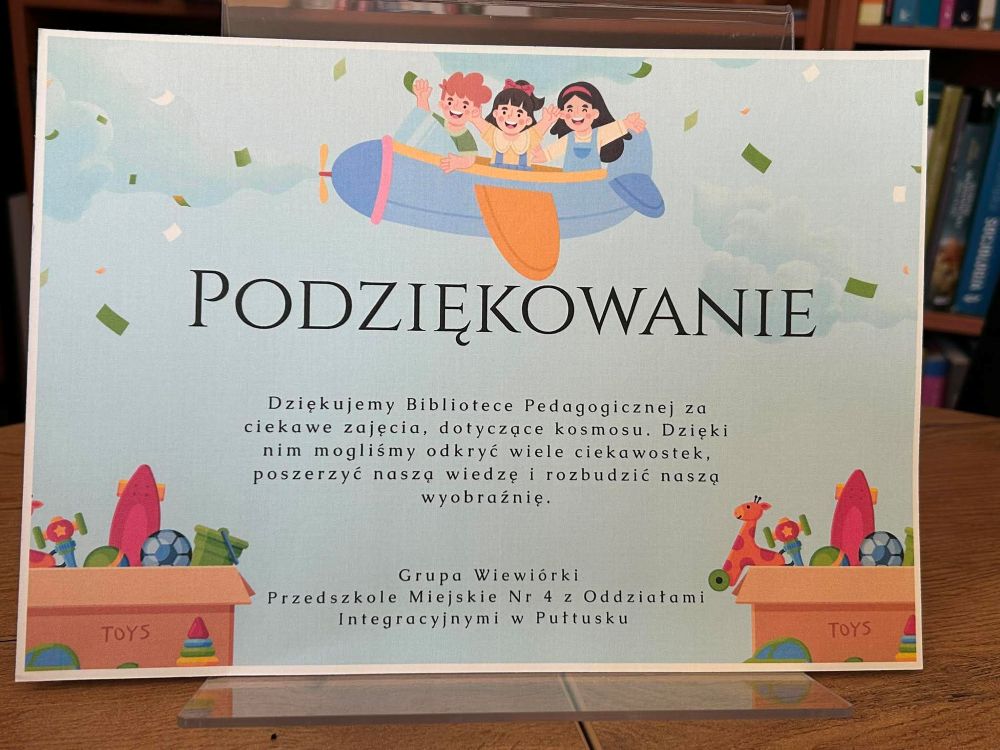 Na zdjęciu widoczne jest estetyczne podziękowanie przygotowane dla Biblioteki Pedagogicznej za przeprowadzenie zajęć o tematyce kosmicznej.
Dyplom utrzymany jest w jasnej, dziecięcej stylistyce. U góry znajduje się ilustracja dzieci lecących samolotem, a całość ozdobiona jest kolorowymi zabawkami i konfetti, co nadaje pracy radosny i przyjazny charakter.
Treść podziękowania podkreśla, że dzięki zajęciom dzieci:
odkryły wiele ciekawostek o kosmosie,
poszerzyły swoją wiedzę,
rozbudziły wyobraźnię i ciekawość świata.
Podziękowanie zostało przygotowane przez grupę „Wiewiórki” z Przedszkola Miejskiego Nr 4 z Oddziałami Integracyjnymi w Pułtusku.
Całość wyraża wdzięczność za inspirujące zajęcia edukacyjne oraz pokazuje, jak ważną rolę pełni biblioteka w rozwijaniu dziecięcych pasji i zainteresowań.