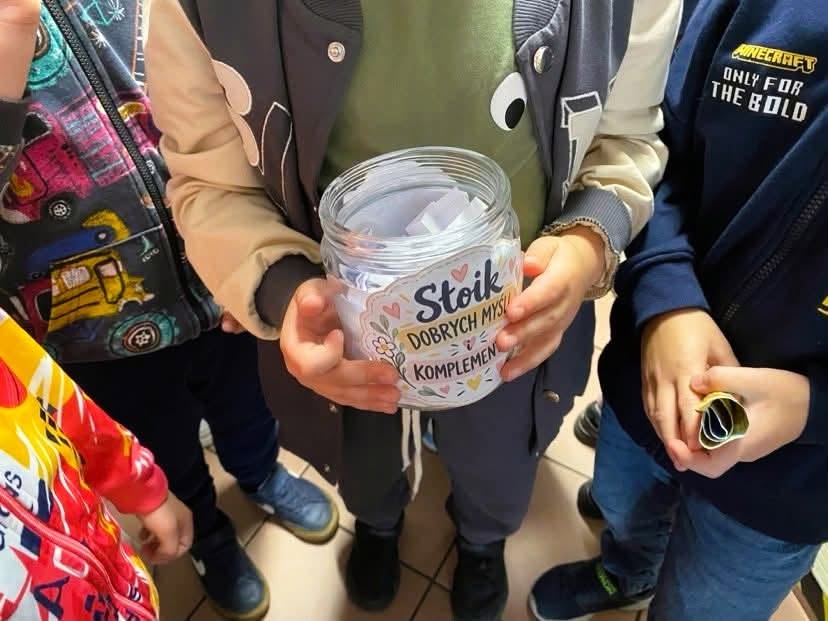 Na zdjęciu widać grupę dzieci stojących wokół przezroczystego pojemnika wypełnionego zwiniętymi karteczkami. Jedno z dzieci trzyma pojemnik oznaczony kolorową etykietą z napisem „Słoik dobrych myśli i komplementów”. Wokół znajdują się inne dzieci w kolorowych bluzach i kurtkach. Fotografia przedstawia aktywność integracyjną lub wychowawczą, zachęcającą do dzielenia się życzliwymi słowami i pozytywnymi wiadomościami.